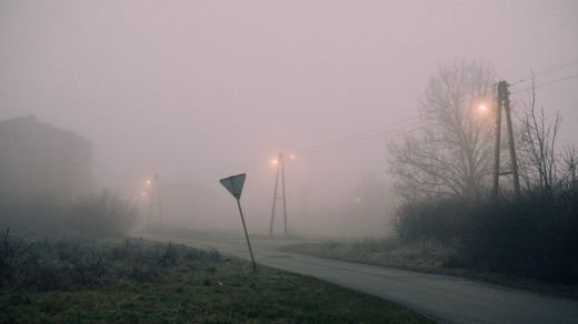 Moody fog envelops a rural street at night, illuminated by street lamps.