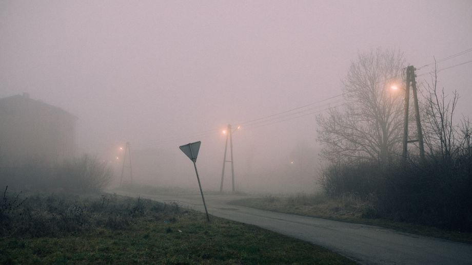 Moody fog envelops a rural street at night, illuminated by street lamps.